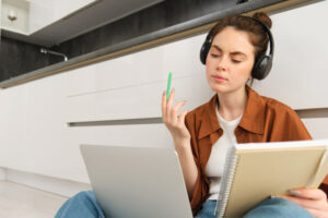 Concentrated young student, woman working on assignment, doing homework on laptop, making notes in notebook during online lesson, remote college course